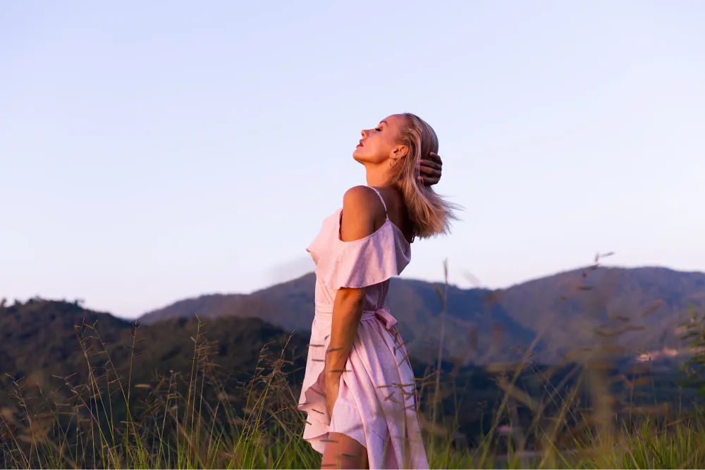Una donna con un abito rosa chiaro è in piedi nell'erba alta, con le montagne sullo sfondo, inclinando la testa all'indietro e toccandosi i capelli.