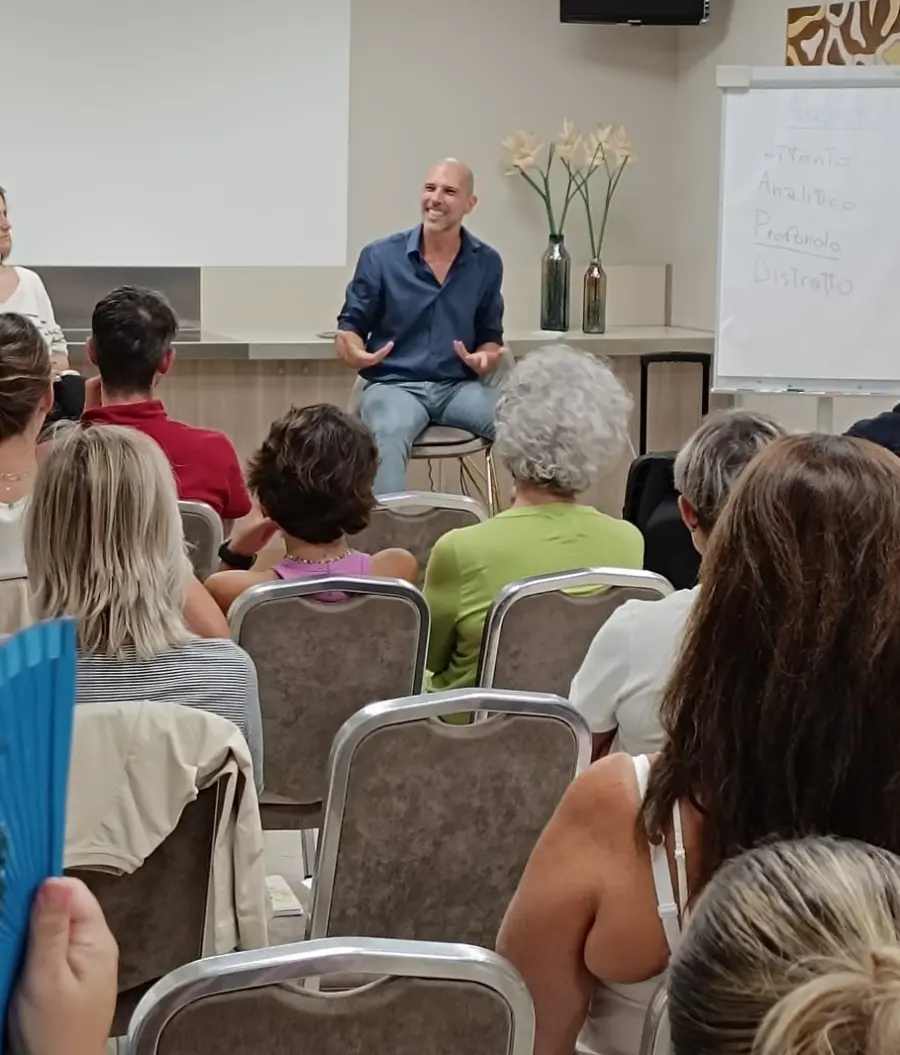 Un uomo sta parlando a un pubblico in un contesto di evento seduto. Sono visibili un cartellone con scritte e vasi con fiori.