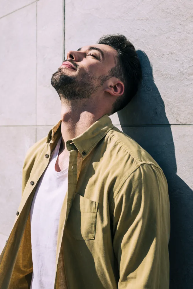 Un uomo con i capelli corti e scuri e la barba è appoggiato a un muro di pietra chiara, con gli occhi chiusi e il viso rivolto verso l'alto, e indossa una maglietta gialla sopra una maglietta bianca.