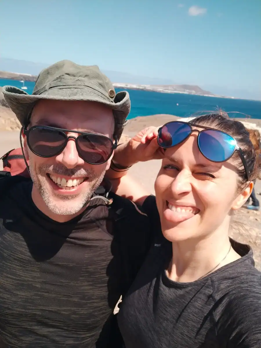 Due persone con occhiali da sole e abiti casual posano per un selfie all'aperto vicino all'oceano in una giornata di sole. Una persona sorride e l'altra tira fuori la lingua in modo giocoso.