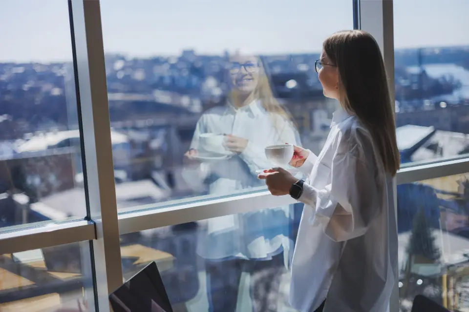 Una donna in camicia bianca &egrave; in piedi accanto a una grande finestra con una tazza di caff&egrave; in mano, con il suo riflesso visibile nel vetro. Sullo sfondo si vede lo skyline della citt&agrave;.