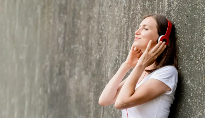 Una donna &egrave; appoggiata a un muro con gli occhi chiusi, indossa delle cuffie rosse e una maglietta bianca.