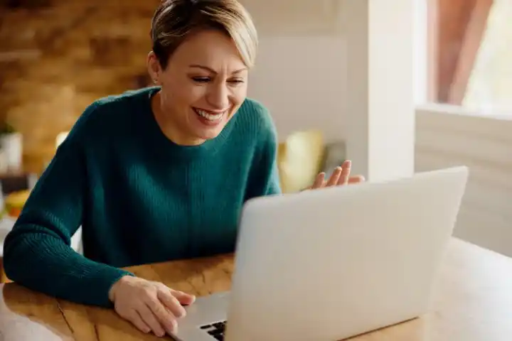 Una persona con i capelli corti e un maglione verde acqua sorride e guarda un computer portatile su un tavolo di legno, salutando lo schermo.