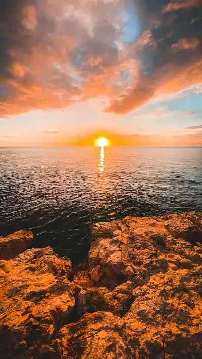 Tramonto su un oceano calmo con scogliere rocciose in primo piano e nuvole illuminate dal sole al tramonto.