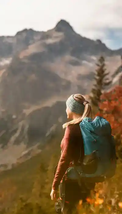 Una persona con zaino e fascia blu si trova all'aperto e guarda la cima di una montagna lontana circondata da alberi e fogliame autunnale.