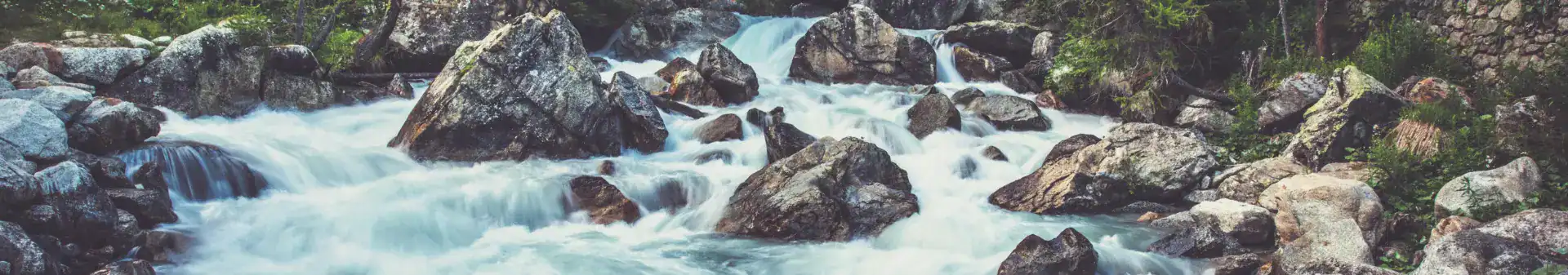 Un torrente di montagna che scorre veloce, con acqua bianca che scorre su grandi rocce, circondato dal verde e da un muro di pietra sulla destra.