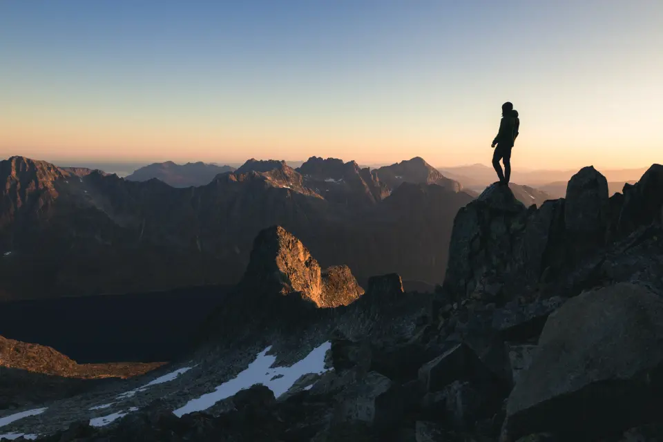 Una persona &egrave; in piedi su una cima rocciosa che domina una catena montuosa al tramonto, con un cielo limpido e un terreno accidentato.